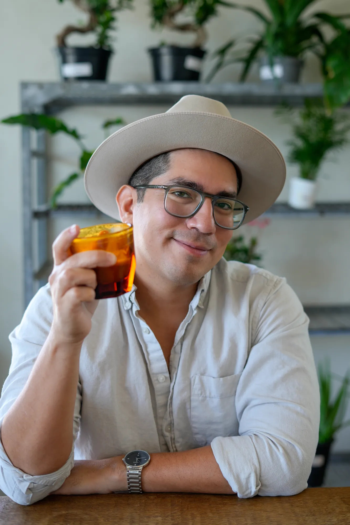 Man in a wide-brim hat raising an iced drink in a plant-filled café